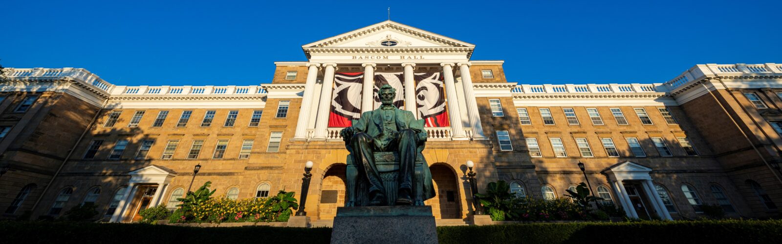 The front of Bascom Hall featuring the statue of Abe Lincoln, a Bucky Badger Banner and a blue sky background